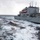 170212-N-HI376-117 Two naval vessels are conducting an underway replenishment (UNREP) operation on rough open seas under overcast skies. A cargo load suspended by a highline transfer system is visible mid-transfer between the two ships. Crew members in safety gear are visible on the deck of the receiving vessel on the left.