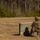 Frag out; Marines participate in grenade assault course A military service member in camouflage uniform and full combat gear is moving tactically alongside a concrete block wall in an open field training area. The image is slightly motion-blurred, suggesting the individual is in rapid movement. A wooded tree line is visible in the background, consistent with a military training range environment.