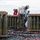 USS America conducts ammo on-load Military personnel are handling large green munitions/bombs secured in wooden crates on what appears to be a naval vessel deck. The ordnance is marked with lot numbers and safety warnings, and multiple service members in protective gear are overseeing or managing the ammunition transfer. Black and red safety straps are scattered across the deck surface.