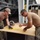 170214-N-N0901-001 Two U.S. Navy personnel in digital camouflage uniforms and tan t-shirts are measuring and marking a sheet of plywood on a makeshift workbench outdoors. They appear to be preparing to cut the wood using a Skilsaw circular saw visible on the board. The setting appears to be a military installation or staging area with heavy equipment and shipping containers in the background.