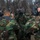 MCESG Gas Chamber A group of military personnel in woodland camouflage uniforms are wearing M40-series gas masks during what appears to be a CBRN (Chemical, Biological, Radiological, Nuclear) training exercise. The individuals are gathered in a close huddle near a concrete structure, suggesting a buddy-check or mask-fitting inspection procedure. The setting appears to be an outdoor military training facility with bare trees visible in the background.