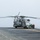 Group Sails A large military helicopter, consistent with a CH-53E Super Stallion, is parked on the flight deck of a naval vessel at sea. Several crew members in flight deck gear are gathered near the aircraft's starboard side, apparently conducting maintenance or pre/post-flight checks. The overcast sky and open ocean horizon confirm an at-sea operational environment.