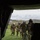 Group Sails The image is taken from inside a military helicopter looking outward through the open rear ramp. Four military personnel in camouflage uniforms and flight helmets are approaching and boarding the aircraft on a green grassy field. The background shows palm trees, rolling hills, and an overcast sky consistent with a California coastal training environment.