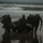 In the CRRC of Time: U.S. Marines and Japanese Soldiers conduct Recon Drills at Exercise Iron Fist A group of military personnel in full tactical gear are carrying a black inflatable rubber raiding craft (IBS/Zodiac-type boat) across a wet beach toward or away from the surf zone. The scene appears to be a combat training exercise or amphibious operation, with rough ocean waves visible in the background. A small vessel is faintly visible on the horizon in the upper left portion of the image.