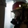 Dwight D. Eisenhower A firefighter or emergency responder is shown in profile view wearing full protective gear in what appears to be a low-visibility or smoky environment. The individual is equipped with a red helmet, face shield, respirator mask, and turnout gear. A mounted headlamp on the helmet is actively illuminated, suggesting operation in a dark or smoke-filled space.