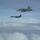 170214-N-XZ522-002 Two military aircraft are flying in formation above a cloud layer against a blue sky. The upper aircraft is a U.S. Navy F/A-18 Hornet or Super Hornet with visible tail number '303', while the lower aircraft appears to be a U.S. Air Force B-1B Lancer strategic bomber. The image appears to document a joint military aviation operation or escort mission.