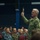 170214-N-DX349-068 A senior U.S. Navy officer in green digital camouflage (NWU Type III) uniform is standing and holding up what appears to be a printed photograph or booklet to an audience of seated military personnel. The setting appears to be a military auditorium or theater, with rows of service members in various Navy uniforms attentively watching. The scene suggests a briefing, all-hands meeting, or training presentation.