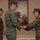 MCIOC Relief and Appointment Three U.S. Marine Corps personnel in MARPAT digital camouflage uniforms are participating in what appears to be a formal military ceremony. A male Marine in the center is exchanging or receiving a ceremonial baton/mace with a female Marine on the right, while a third Marine stands at attention in the background. The setting appears to be an indoor ceremonial hall with a framed painting visible on the wall behind them.