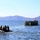 Western Pacific Deployment 2017 Military personnel are conducting a waterborne operation involving two vessels: a black inflatable rubber raiding craft (CRRC) carrying several soldiers in combat gear, and a larger amphibious assault vehicle or floating platform flying a United States flag. The scene appears to be a military exercise or amphibious landing operation taking place in a calm bay or coastal waterway with mountainous terrain visible in the background. The presence of the American flag and military equipment suggests a U.S. military or joint allied forces training exercise.