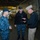 Capt David O. Bynum, chaplain, U.S. Pacific Fleet, tours USS Bonhomme Richard (LHD 6) Three U.S. Navy personnel are engaged in conversation inside what appears to be the well deck or hangar bay of a naval vessel. Two individuals are wearing Navy Working Uniform (NWU) digital camouflage, while the third wears a combination cover (white cap) and khaki trousers indicating senior officer rank. The setting is a large interior ship compartment with industrial infrastructure visible in the background.