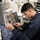 USS America conducts replenishment at sea with USNS Henry J. Kaiser A young man in dark navy coveralls is working on a mechanical or hydraulic component featuring copper tubing and fittings, labeled 'MU59' or similar. He appears to be in a technical workspace or workshop environment with desks, paperwork, and filing trays visible in the background. His focused expression and attire suggest a professional or military technical maintenance context.