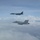 170214-N-XZ522-003 Two U.S. military aircraft are flying in formation above a cloud layer under a partly cloudy sky. The formation consists of a large strategic bomber in the upper portion of the frame and a smaller tactical fighter jet in the lower portion. Both aircraft appear to be on a joint mission or escort flight.