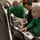 Dwight D. Eisenhower Three individuals wearing green shirts are working together on electronic or mechanical equipment in what appears to be a military or industrial hangar environment. Two of the individuals wear green long-sleeve shirts with insignia patches, suggesting military affiliation, while one uses a multimeter and another operates a ruggedized laptop. They appear to be performing diagnostics or maintenance on a piece of equipment.