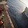 170214-N-NJ910-0089 A naval replenishment-at-sea (RAS) operation is underway between two military vessels sailing in close parallel formation on open ocean. Several crew members wearing safety helmets and orange life vests are positioned on the deck of one ship, managing equipment and lines. The image is taken from an elevated angle, likely from a superstructure or mast, showing the gap between the two ships and the wake generated by their movement.