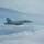 170214-N-XZ522-001 A U.S. Navy F/A-18 Hornet or Super Hornet jet fighter, marked with the modex number '303', is photographed in flight above a cloud layer. The aircraft appears to be in level cruise flight and is photographed from another aircraft flying in close proximity. The image shows the aircraft in standard gray naval aviation livery with a squadron insignia visible on the tail.