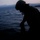 RIMPAC’s lost shots A silhouetted figure wearing military-style tactical gear, including a helmet and what appears to be body armor with a communications headset, is crouched near the water's edge at dusk or dawn. The figure is positioned against a body of water with mountainous terrain visible in the background. The low-light conditions render the subject almost entirely in silhouette.