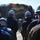 170214-N-YG414-136 A military officer in Navy Working Uniform (NWU) addresses a group of personnel wearing blue hard hats in an outdoor setting. The gathering appears to be a safety briefing or operational muster, with a rocky cliff face visible in the background. A larger formation of similarly equipped personnel is visible further in the background.