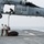 USS America conducts ammo on-load A U.S. Navy MH-60S Seahawk helicopter (number 61, assigned to HSC-21) hovers low over a ship's flight deck while two deck crew members use a grounding pole to discharge static electricity before attaching a cargo load. A crew member is visible seated in the open cabin door of the helicopter. The scene appears to be taking place at sea under overcast conditions.