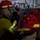 Dwight D. Eisenhower The image shows personnel in firefighting or damage control gear, wearing red helmets with yellow markings and breathing apparatus, inside what appears to be a naval vessel or industrial facility. A person in the background stands in a watertight doorway holding a sign with the letter 'B', likely indicating a training or drill scenario. The setting features metal bulkheads, electrical panels, pipes, and a characteristic oval watertight hatch consistent with a military ship.