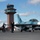 170206-N-IM832-068 A military airfield scene showing a distinctively painted teal/blue camouflage F-16 Fighting Falcon aircraft being marshaled by ground crew personnel on the tarmac. The aircraft bears the marking 'NAWDC' (Naval Aviation Warfighting Development Center) on its fuselage, indicating it is an adversary/aggressor aircraft. An air traffic control tower and hangar facilities are visible in the background.