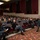 170215-N-JD834-006 A large group of U.S. Navy personnel in Navy Working Uniform (NWU) digital camouflage are seated in an auditorium or theater setting. A senior officer or official is standing at the front-right of the audience, appearing to address or interact with the assembled sailors. The venue features red/maroon theater seating, a patterned carpet, and dark acoustic panels on the walls.