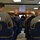 Inspector gives brief A briefing or meeting is taking place in what appears to be a military conference room, with personnel seated in chairs bearing 'INSURV' patches and 'USS Nimitz CVA-68' insignia. A presenter stands at the front of the room holding papers, with a projection screen visible behind him. The attendees are wearing military uniforms including Navy Working Uniform (NWU) and what appear to be flight suits or coveralls.