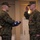 MCIOC Relief and Appointment Two U.S. Marines in MARPAT digital camouflage uniforms are participating in what appears to be a formal military ceremony, likely a retirement or award ceremony. The Marine on the right is rendering a salute and wearing a medal with a red ribbon, while the Marine on the left holds a folded American flag. The indoor setting with neutral-toned walls suggests a banquet hall or ceremonial venue on a military installation.