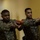 Provost Marshal's Office active shooter training Two U.S. Marines in MARPAT camouflage uniforms are conducting a close-quarters battle (CQB) or room-clearing training exercise in an indoor corridor. They are holding orange/red training pistols (inert dummy weapons used for drills) in an extended shooting stance. Additional Marines in uniform are visible in the background observing or waiting their turn.