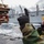 170212-N-HI376-334 A naval service member wearing a yellow hard hat, camouflage uniform, and black gloves is directing operations on the deck of a military vessel at sea. They appear to be signaling with one hand raised, likely coordinating a replenishment-at-sea (RAS) or cargo transfer operation. A large gray military supply ship is visible in the background alongside stacked wooden pallets on the deck.