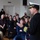 170209-N-VK873-265 A naval officer in dress uniform is addressing a large group of young people, likely students, in what appears to be a school or community hall. The officer, wearing a white-topped cap and dark uniform with gold sleeve stripes, is gesturing with his hands while speaking. The audience, consisting of approximately 30-40 individuals, is seated and attentively listening.