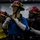 Dwight D. Eisenhower Four individuals wearing firefighting or damage control gear, including red helmets with face shields, protective hoods, and gloves, are engaged in what appears to be a training drill or emergency response exercise. The person in the center is holding a fire hose nozzle while the others support from behind in a hose team formation. The setting appears to be an indoor facility, possibly aboard a naval vessel based on the equipment and attire.