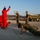 KMEP 17-8 A group of military personnel in olive/camouflage uniforms and individuals in red tracksuits are engaged in physical training exercises on a paved outdoor surface. Some participants are doing push-ups while others appear to be cheering or encouraging them. A camouflage-painted military truck is visible in the background, suggesting this is a military base or training facility.