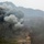 KMEP 17-8 A large plume of dark gray and white smoke rises from a mountainous, forested hillside. The terrain appears to be in late autumn or winter, with bare deciduous trees and sparse evergreen vegetation. A small structure or vehicle is faintly visible at the base of the smoke column near a dirt road.