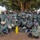 170213-N-N0901-001 A large group of military personnel, approximately 30-40 individuals, posed for a group photograph under a large tree in what appears to be a rural outdoor setting. The majority wear matching camouflage uniforms with tan/beige helmets and body armor vests, with visible Uganda People's Defence Force (UPDF) patches. One individual in the back row appears to be wearing a different uniform style, possibly a foreign advisor or trainer.