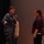 170214-N-XF155-180 A U.S. Navy service member in blue digital camouflage (NWU Type I) uniform is holding a microphone and speaking to a civilian woman in a plaid shirt on what appears to be a stage or auditorium setting. A third person in a tan/beige cardigan is partially visible in the background. The scene suggests a public event, Q&A session, or community outreach engagement.