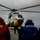 RIMPAC’s lost shots A military or naval helicopter, appearing to be a gray medium-lift rotary-wing aircraft (likely a Mil Mi-8 or similar), is on the flight deck of a ship with its rotors spinning. Three deck crew members wearing colored safety vests and hearing protection are observing or directing the helicopter operation. The scene takes place under overcast skies with open sea visible in the background.
