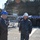 170214-N-YG414-130 A military official wearing a white hard hat labeled 'DALTON' in Navy Working Uniform (NWU) is engaged in conversation with another person in a black coverall and blue hard hat, likely on a naval vessel deck or shipyard. A large formation of sailors wearing blue hard hats and dark uniforms is assembled in the background. The setting appears to be an outdoor naval facility, possibly in Japan based on the hillside vegetation.