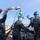 170215-N-KP948-045 A group of U.S. Navy sailors in digital camouflage uniforms (NWU Type I) are working on the deck of a naval vessel, handling what appears to be a torpedo or similar cylindrical ordnance/equipment using a crane or davit system. The operation is taking place in what appears to be a naval shipyard or port facility, visible in the background. Multiple personnel are wearing blue hard hats, indicating a safety-regulated work environment.