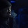 Hué City, part of the George H.W. Bush Carrier Strike Group (GHWBCSG), is conducting naval operations in the U.S. 6th Fleet area of operations in support of U.S. national security interests A close-up portrait of a man illuminated by a single directional light source against a dark, blue-tinted background. He is wearing a black knit beanie and a dark jacket with a velcro patch visible on the collar. The dramatic low-key lighting creates strong contrast, highlighting one side of his face while the other remains in shadow.