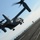 170214-N-NJ910-0248 A military tiltrotor aircraft, identifiable as a Bell Boeing V-22 Osprey, is captured in low-altitude flight over what appears to be the flight deck of an aircraft carrier at sea. The rotors are in a transitional or helicopter-mode position, with significant motion blur indicating active rotation. A flight deck crew member in a yellow vest is visible in the background on the carrier deck.