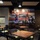 Starbucks Grand Opening The image shows the interior of a Starbucks coffee shop located at Camp Joseph H. Pendleton, a U.S. Marine Corps base in California. The seating area features dark wood tables and chairs with green cushions, arranged on a tiled floor. A large decorative mural styled as a vintage postcard reading 'Greetings from Camp Joseph H. Pendleton, California' dominates the back wall, featuring military imagery and a uniformed Marine figure.