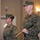 MCIOC Relief and Appointment Two U.S. Marines in MARPAT woodland camouflage uniforms are standing at attention in what appears to be an indoor ceremonial setting. The male Marine in the foreground is holding a ceremonial NCO sword with a gold handle, suggesting a formal military ceremony or event. A female Marine stands at ease in the background.