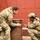 170213-N-N0901-008 Two individuals in military-style multicam camouflage uniforms are crouched near a metal barrel against a red tiled wall. One person is wearing full tactical gear including a helmet with communications equipment and body armor, while the other is in camouflage clothing without a helmet. They appear to be engaged in a close consultation or tactical discussion over the barrel.