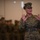 MCIOC Relief and Appointment A female U.S. Marine in MARPAT digital camouflage uniform is speaking or instructing with both hands raised, palms facing outward. Her name tape reads 'RUIZ' and her chest tape reads 'U.S. MARINES'. In the blurred background, multiple uniformed personnel are visible, suggesting a formation or briefing setting.