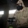 MCESG Gas Chamber A person wearing desert camouflage military uniform, a gas mask, and protective goggles is operating a portable camp stove inside a dimly lit building. Smoke or vapor is visibly rising from a pan or container on the stove. A second individual in similar protective gear is visible in the background near a window.