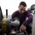 Sailor maintains equipment A young man in a purple long-sleeve shirt is performing maintenance or repair work on industrial gas equipment. He is using a hand tool, likely a wrench or ratchet, to work on components of what appears to be an LP gas service unit. The setting appears to be an outdoor or semi-outdoor industrial/mechanical environment.