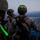 RIMPAC’s lost shots Three military personnel in combat gear are positioned on the deck of a vessel at dusk or dawn, overlooking open water. They appear to be in a briefing or coordination posture, with one individual facing the others and speaking. The scene suggests a maritime operation or amphibious exercise.