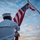 170211-N-BL637-032 A U.S. Navy sailor in dress white uniform is photographed from behind while raising or holding the American flag on a flagpole during what appears to be a sunrise or sunset ceremony. The dramatic sky with scattered clouds and warm golden light creates a striking patriotic composition. The low-angle perspective emphasizes both the sailor and the unfurled flag against the sky.