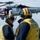 USS America conducts ammo on-load Two U.S. Navy flight deck crew members in yellow vests and helmets stand on the flight deck of what appears to be USS America (LHA-6), communicating while an MH-60 Seahawk helicopter hovers nearby. The helicopter bears the number '70' and 'NAVY' markings, with an American flag painted on its nose. The scene depicts routine flight deck operations aboard a naval vessel at sea.