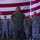 Lt. Gen. Nicholson recognizes Airmen’s support in MV-22 incident A military briefing or address is taking place indoors in front of a large American flag. A senior military figure in Marine Corps MARPAT (woodland digital camouflage) stands with his back to the camera, addressing a group of personnel wearing U.S. Air Force Airman Battle Uniforms (ABU). The setting appears to be a hangar or large military facility.